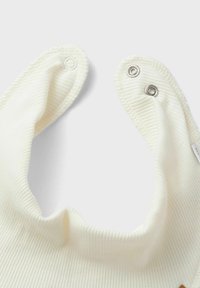 Weißes, geripptes Stofflätzchen mit einem runden Halsausschnitt, das zwei Druckknopfverschlüsse an den Seiten zum Schließen hat. Weicher Stoff, einfaches Design.