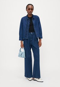 Denim jacket with ruffled detail, paired with high-waisted denim trousers. Black ribbed top, blue handbag, and black-and-white shoes.