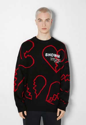 Pull noir avec motifs abstraits de cœur et visage rouges. Texture tricotée, poignets et col côtelés, avec le logo "SNDWN" en blanc sur la poitrine.