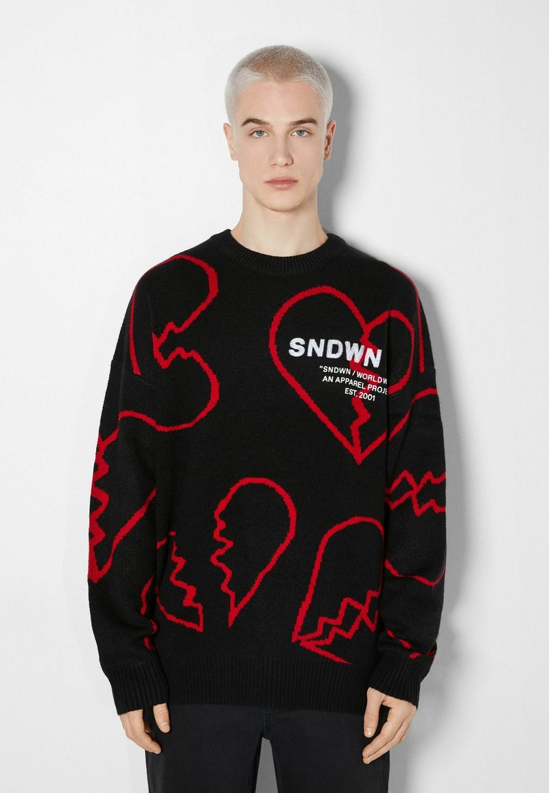 Pull noir avec motifs abstraits de cœur et visage rouges. Texture tricotée, poignets et col côtelés, avec le logo "SNDWN" en blanc sur la poitrine.