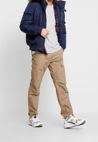 Marineblau puffer Jacke über einem grauen T-Shirt, kariertes Hemd darunter, khakifarbene Cargo-Hosen mit mehreren Taschen und graue sowie weiße Sportschuhe.