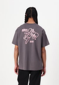 Pilkas medvilninis marškinėliai su dideliu rožiniu įvaizdžiu, kuriame angelas laiko širdį, ir užrašu "ADIDAS ORIGINALS" ant nugaros. Atsipalaidavęs siluetas.