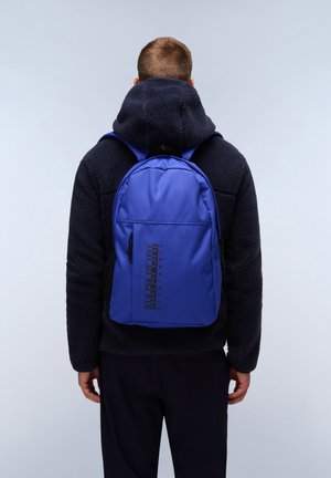 Mochila azul hecha de un tejido duradero, con un bolsillo frontal con cremallera, logo negro y correas ajustables. Llevada sobre una sudadera con forro polar negra.