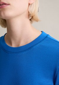 Niebieski top z żebrowanym wzorem i szerokim dekoltem. Faktura jest gładka, a kolor to żywy odcień niebieskiego. Minimalne detale przeszyć.