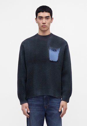 Giovane uomo con capelli corti e neri che indossa un maglione lavorato a maglia scuro con tasca blu e jeans blu, in piedi su sfondo bianco uniforme.