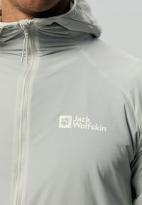 Lichtgrijze jas gemaakt van glad, weerbestendig materiaal, met een capuchon, een rits aan de voorkant en een wit logo op de borst.