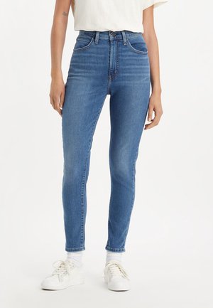 Frau trägt hoch taillierte blaue Skinny-Jeans mit weißen Socken, weißen Sneakers und einem in die Hose gesteckten weißen kurzärmligen Oberteil.