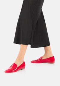 Rote Lackleder-Loafers mit elegantem Design, ausgestattet mit einer leicht erhöhten Zehenkappe und niedrigem Profil. Getragen mit einem schwarzen, fließenden Kleidungsstück.