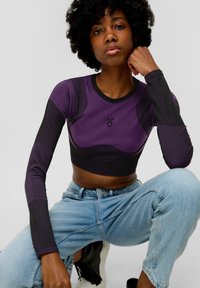 Fioletowy i czarny crop top z długim rękawem, dopasowany fason, prążkowana tekstura i szczegóły logo na klatce piersiowej, zestawiony z jasnoniebieskimi dżinsami.