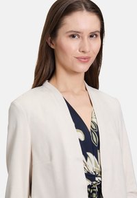 Beżowa marynarka wykonana z miękkiego materiału z kołnierzem typu szal. Nosi ją osoba w wzorzystej, ciemnej sukience z kwiecistymi motywami i knotenym detalem z przodu.