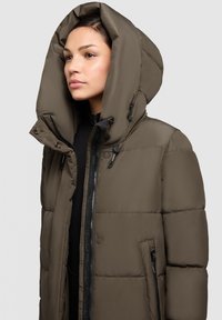 Kūnu jaka izsmalcinātā brūnā krāsā ar lielu kapuci, quiltēta tekstūra, priekšējā rāvējslēdzēja aizdare un sānu kabatas ar melniem aksesuāriem.