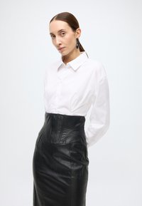 Camisa branca de botões com mangas compridas combinada com uma saia preta de couro de cintura alta, com detalhes texturizados e silhueta ajustada.