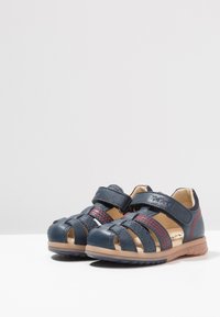 Sandalias infantiles de cuero azul con diseño abierto, correa de velcro, detalles de costura roja y suela de goma marrón texturizada.