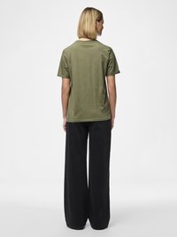 Žena s ravnom plavom kosom, odjevena u maslinasto zelenu majicu s kratkim rukavima i široke crne hlače, okrenuta leđima na bijeloj pozadini.