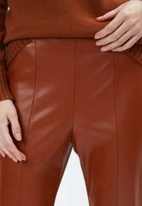 Pantaloni in pelle marrone con una texture liscia, cuciture verticali e un design aderente, abbinati a un maglione lavorato a maglia color ruggine.