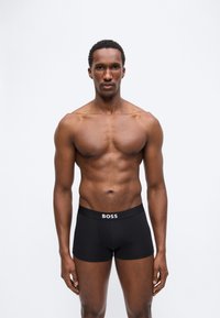 Μυώδης άνδρας που στέκεται με το πρόσωπο προς τα εμπρός, φορώντας μαύρα μπόξερ με την επιγραφή "BOSS", πάνω σε απλό ανοιχτόχρωμο φόντο.