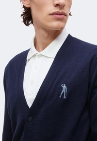 Homme portant un polo blanc sous un cardigan bleu marine avec une petite figure brodée sur la poitrine gauche, montré du milieu du visage jusqu’au torse.