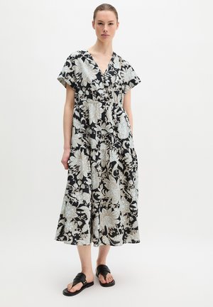 Femme portant une robe midi noire avec un grand imprimé floral blanc, manches courtes, col en V et sandales plates noires, debout devant un fond blanc.