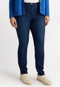 Jeans skinny de tiro alto en azul oscuro, con una textura de mezclilla suave, que cuentan con un cierre frontal de botón y un diseño minimalista. Combinados con zapatillas crema.