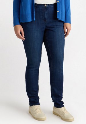 Dunkelblaue, hochtaillierte Skinny-Jeans mit glatter Denim-Oberfläche, ausgestattet mit einem vorderen Knopfverschluss und minimalistischem Design. Kombiniert mit cremefarbenen Sneakers.