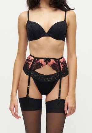 Femme portant un soutien-gorge en dentelle noire, une culotte assortie, un porte-jarretelles en dentelle florale rose et noire, et des bas noirs jusqu'aux cuisses sur un fond uni.