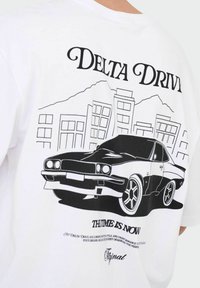 Biała bawełniana koszulka z czarnym nadrukiem przedstawiającym samochód i panoramę miasta. Pod grafiką znajduje się napis "DELTA DRIVE" oraz "THE TIME IS NOW".