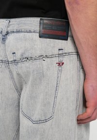 Koptatott világoskék farmernadrág látható varrási részletekkel, texturált hátlap címkével és piros díszítő varrással a zseb közelében.