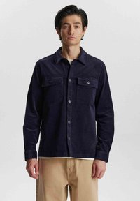 Cămașă din corduroy bleumarin cu guler, două buzunare pe piept și închidere cu butoane. Textură moale și mâneci lungi, combinată cu pantaloni bej.