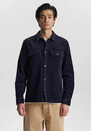 Cămașă din corduroy bleumarin cu guler, două buzunare pe piept și închidere cu butoane. Textură moale și mâneci lungi, combinată cu pantaloni bej.