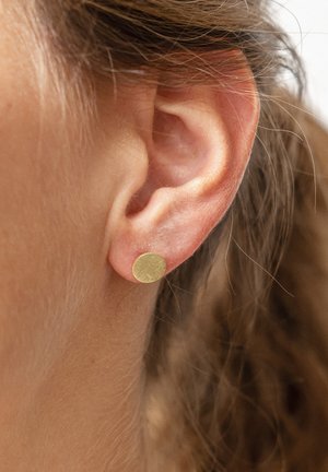 Primo piano di un orecchio sinistro che indossa un piccolo orecchino a bottone rotondo in oro con capelli castano chiaro raccolti all'indietro.