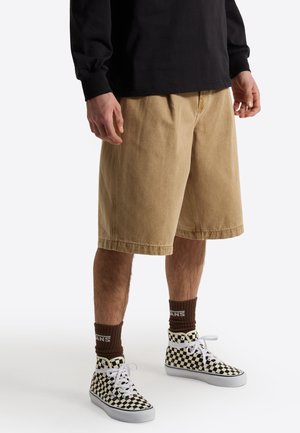 Persoon draagt beige kniebroek, zwart shirt met lange mouwen, bruine Vans-sokken en zwart-witte geruite Vans-sneakers.