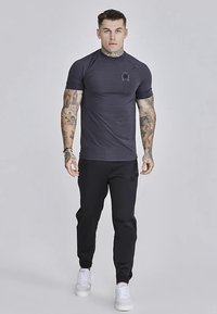 Tummanharmaa lyhythihainen urheilupaita, jossa teksturoitu kangas ja pieni vaakuna rinnassa, yhdistettynä mustiin housuihin ja valkoisiin lenkkareihin.