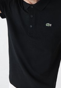 Polo noir en coton, avec un col côtelé, une patte à trois boutons et un logo crocodile vert brodé sur la poitrine.
