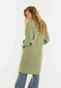 Vrouw met lang krullend haar, gekleed in een groene gebreide cardigan met zakken en blauwe spijkerbroeken, glimlachend en naar beneden kijkend.