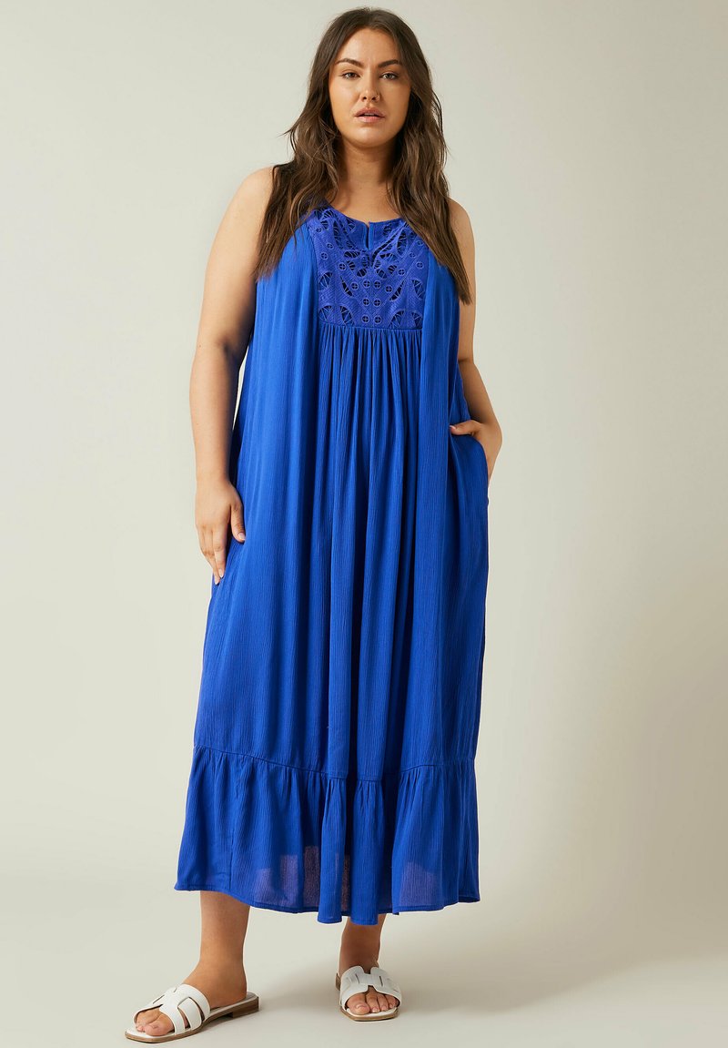 Blaues Maxikleid mit einem strukturierten Oberteil mit ausgeschnittenen Designs, lockerer Passform, geraffter Taille und rüschenbesäumtem Saum. Getragen mit weißen Sandalen.