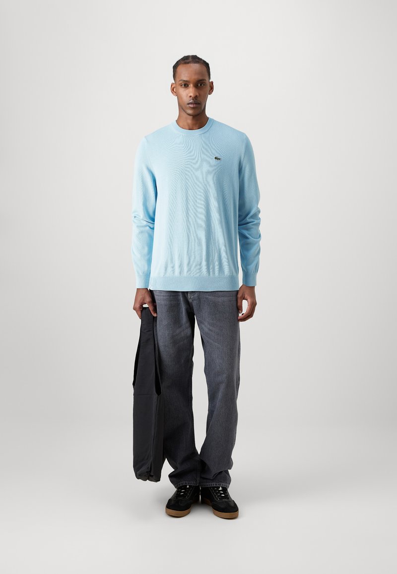 Pull en laine léger bleu clair avec un col rond et des poignets côtelés ; associé à un jean en denim gris foncé et des baskets noires, tenant un sac noir.