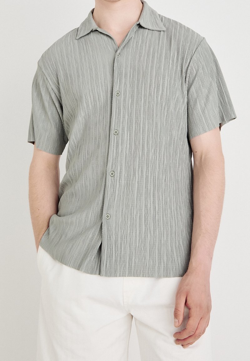 Camicia a maniche corte con bottoni di colore grigio chiaro, impreziosita da righe verticali testurizzate e da un colletto morbido. Presenta bottoni chiari sul davanti.