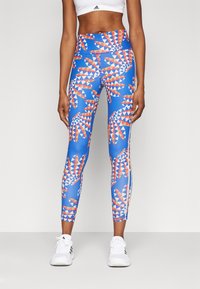 Leggings azuis até ao tornozelo com um padrão geométrico vermelho e branco, apresentando uma cintura alta e combinados com sapatilhas brancas.