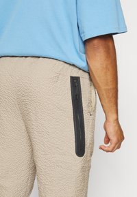 Beige byxor med struktur och en svart ficka med dragkedja. Bärs med en ljusblå kortärmad skjorta som visar en del av armen.