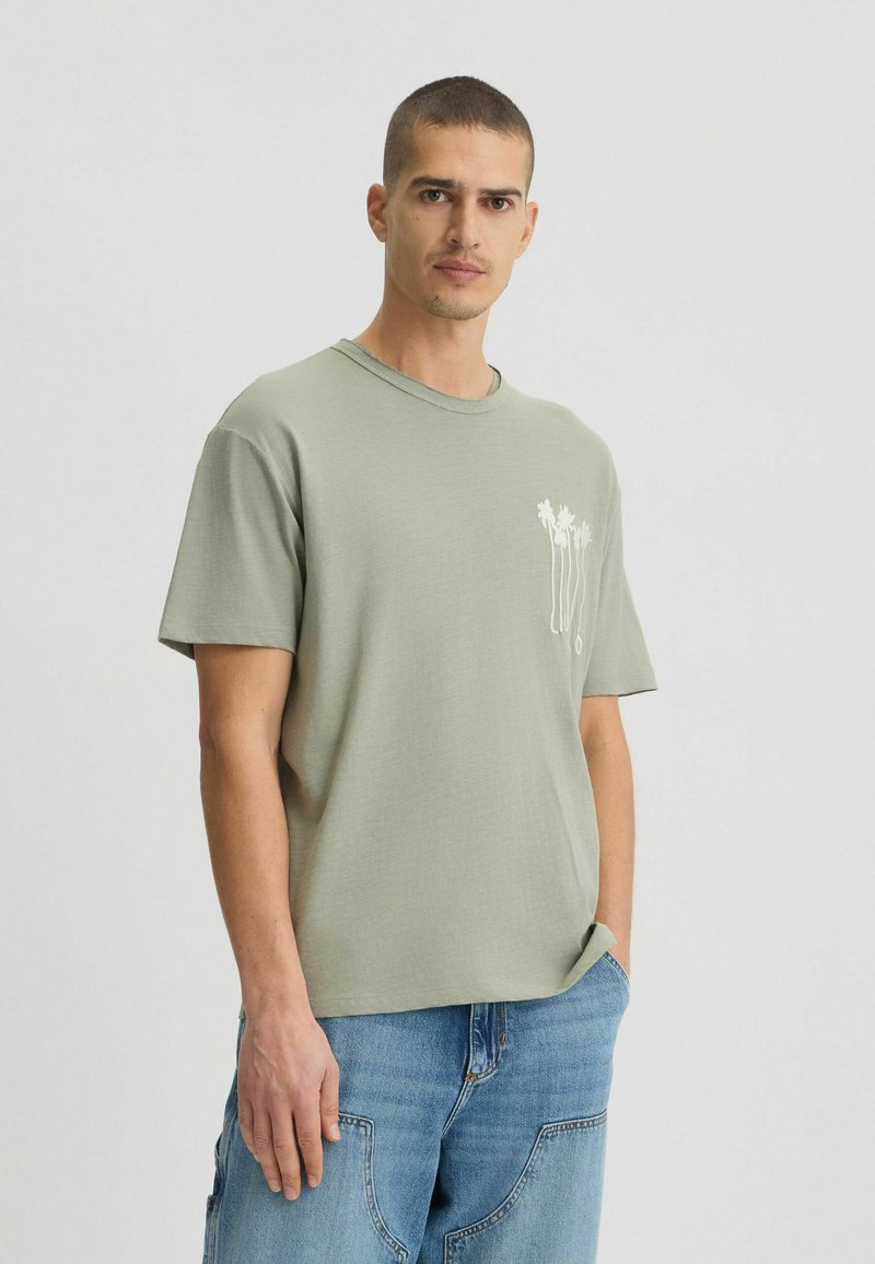 Rövid ujjú, zsálya zöld póló, puha anyagból készült, bal oldalon fehér pálmafával. Laid-back fit és kerek nyakú.