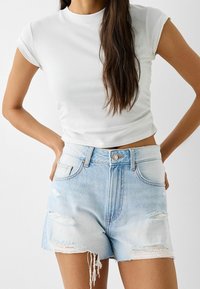 T-shirt blanca recortada de manga corta combinada con pantalones cortos de mezclilla de color azul claro con detalles desgastados y dobladillo deshilachado.