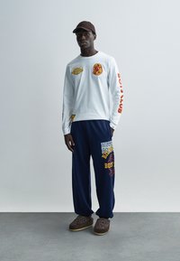 Πουκάμισο λευκό με μακριά μανίκια και τυπωμένα γραφικά, σε συνδυασμό με μπλε σκούρο φόρμες με έντονο κείμενο και σχέδια. Καφέ παπούτσια τύπου slip-on ολοκληρώνουν την εμφάνιση.