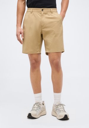 Άνδρας που φοράει μπεζ σορτς μήκους μέχρι το γόνατο, λευκές κάλτσες και μπεζ αθλητικά παπούτσια, στέκεται με το ένα χέρι στην τσέπη σε ουδέτερο φόντο.