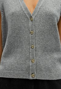 Gilet tricoté gris avec un profond décolleté en V. Détails de boutons en forme de nœud dorés sur le devant et un ourlet en côtes. Texture douce et manches courtes.