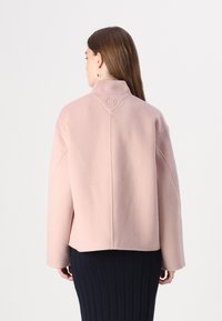 Rosa Wolljacke mit hohem Kragen, fallenden Schultern und einem hexagonalen Logodetail am Hals, kombiniert mit einem gerippten marineblauen Rock.