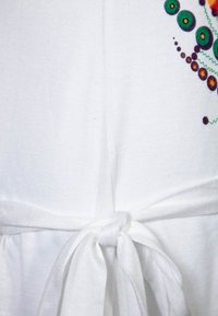 Vit klänning i tyg med ett bälte i midjan. Har färgglad brodyr på sidan, inklusive gröna cirklar och svängande mönster.