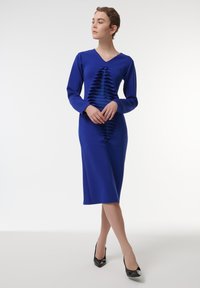 Vestido azul de manga larga con escote en V, que presenta recortes horizontales en capas a lo largo del centro y una silueta ajustada, combinado con tacones negros.