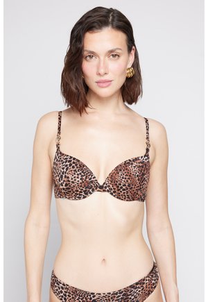 Vrouw met donker haar op schouderlengte, die gouden oorringen draagt en een beha met luipaardprint en bijpassende slip, voor zich uitkijkend tegen een witte achtergrond.