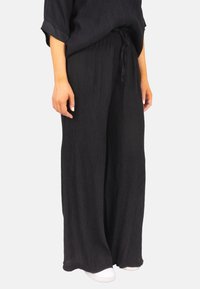 Pantalons larges noirs en tissu texturé, avec une taille élastique et un détail de cordon de serrage. Portés avec des chaussures blanches pour le contraste.