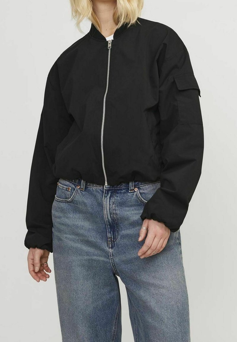 Personne portant un blouson bomber noir zippé avec une poche sur la manche et un jean taille haute bleu clair.